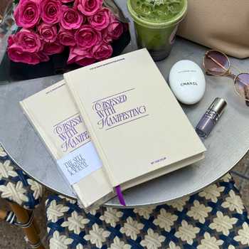 Two books titled 'Obsessed with Manifesting' on a table with pink roses, a green smoothie, a Chanel earring, and sunglasses.
