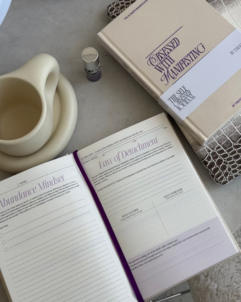Open notebook with pages titled 'Law of Detachment' and 'Abundance Mindset', next to a closed book titled 'Obsessed with Manifesting'.