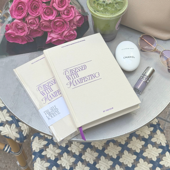 Two books titled 'Obsessed with Manifesting' on a table with pink roses, a green drink, sunglasses, and a Chanel bottle.